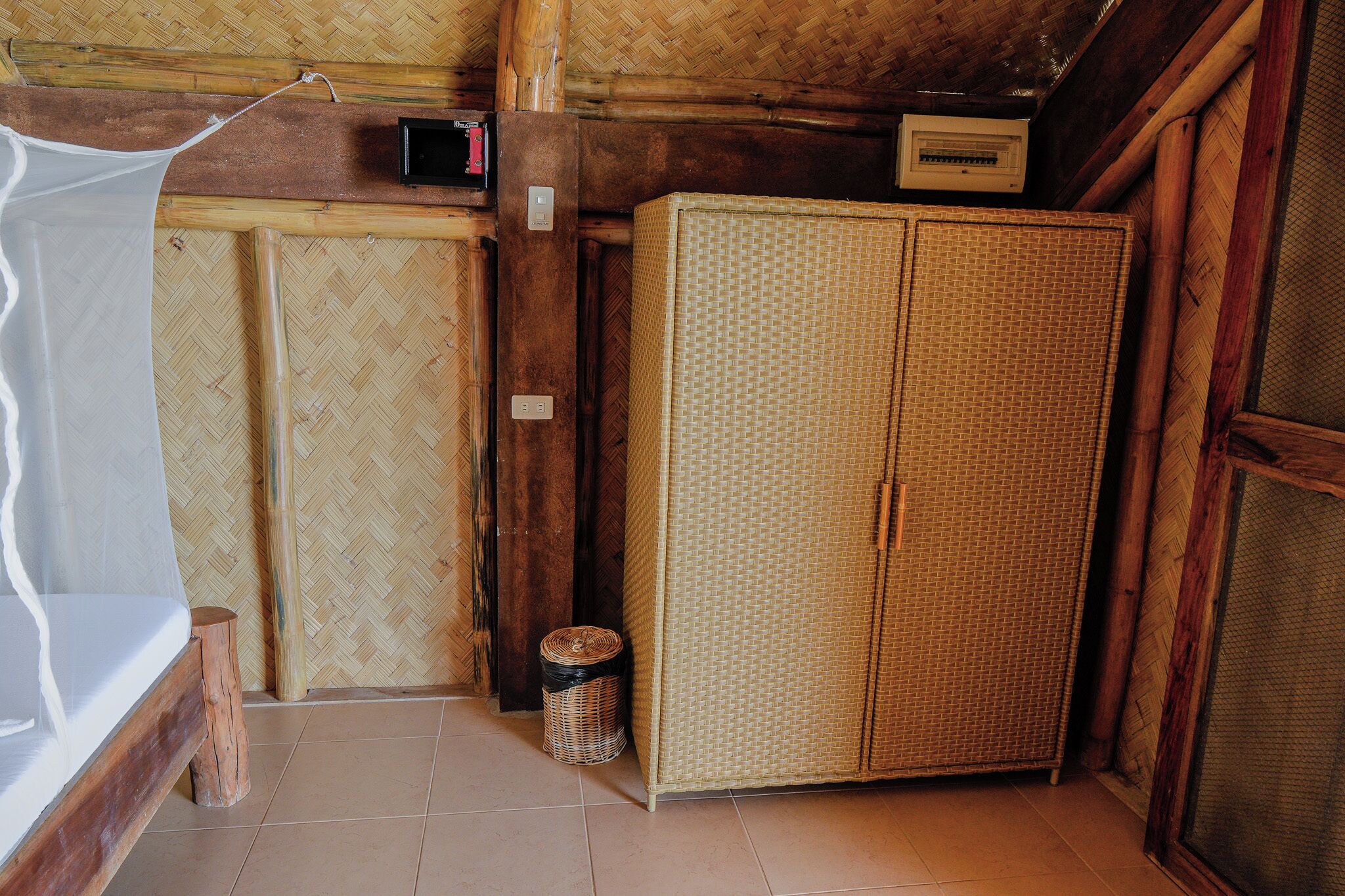 A room with a bed, a wicker wardrobe, and a small trash bin.