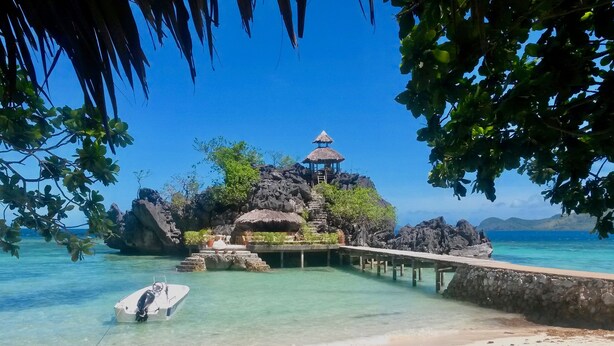 A tropical beach with a thatched-roof structure on a rocky outcrop, a wooden pier leading to a boat, and clear turquoise waters. - Sangat Island Dive Resort (Coron)