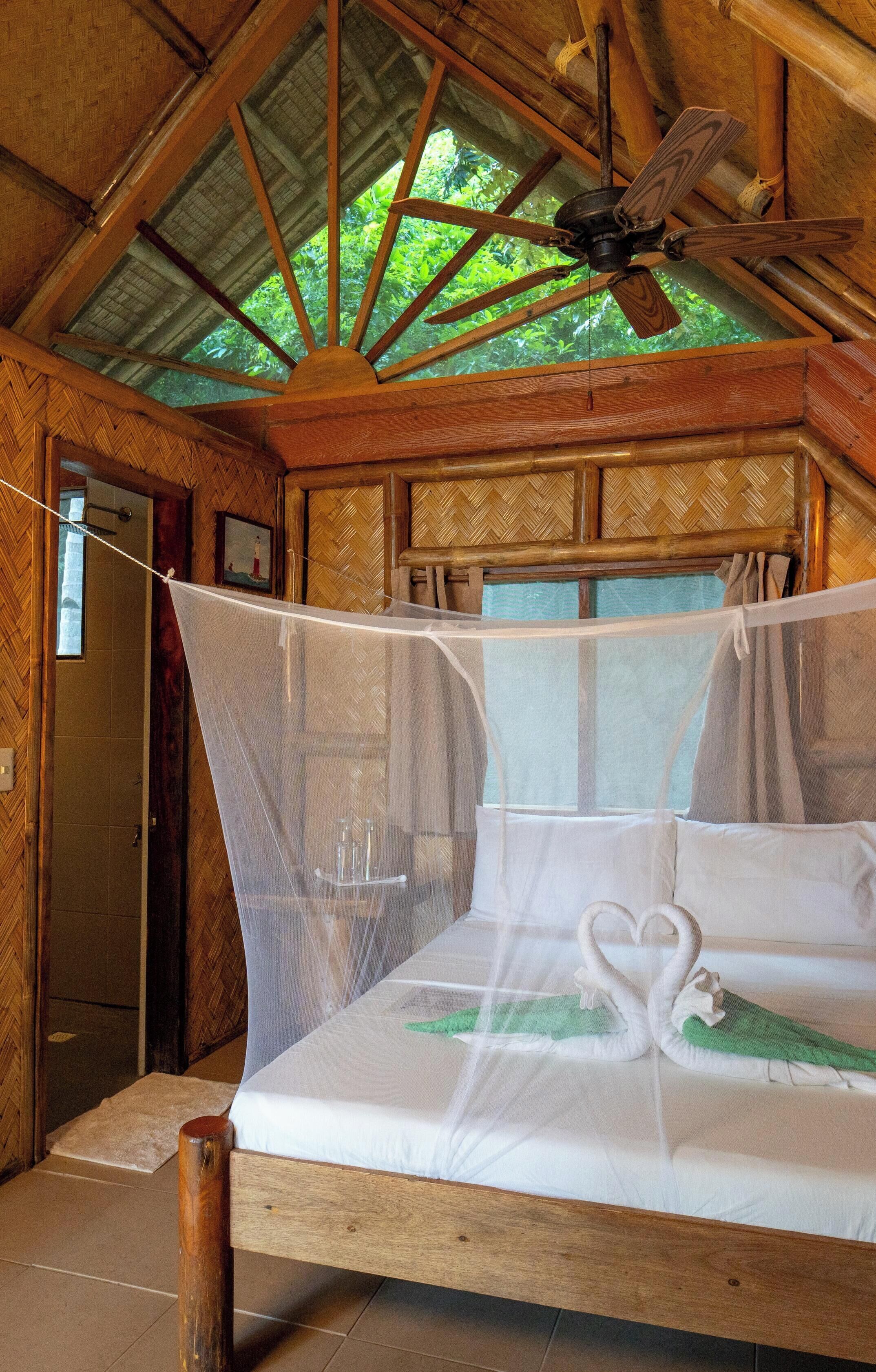 Quarto de madeira com uma cama, um ventilador de teto e uma janela com vista para vegetação.