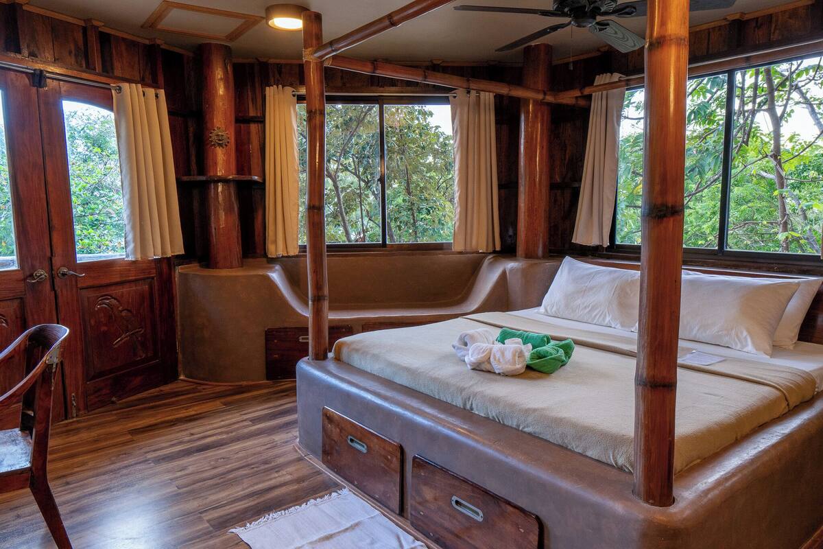 A four-poster bed with a canopy, wooden walls, and large windows offering a view of trees.