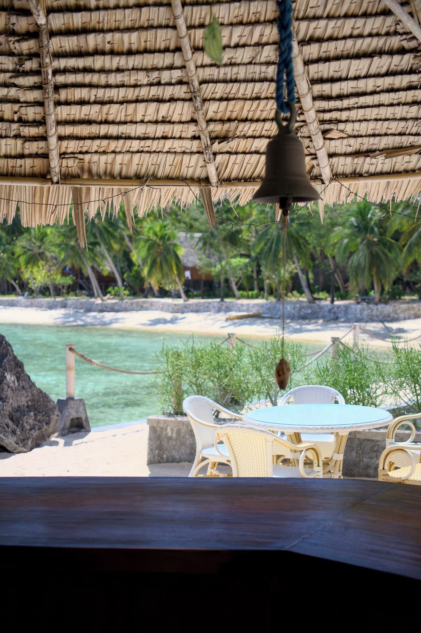 A beachside dining area with a thatched roof, wicker chairs, and a hanging bell.
