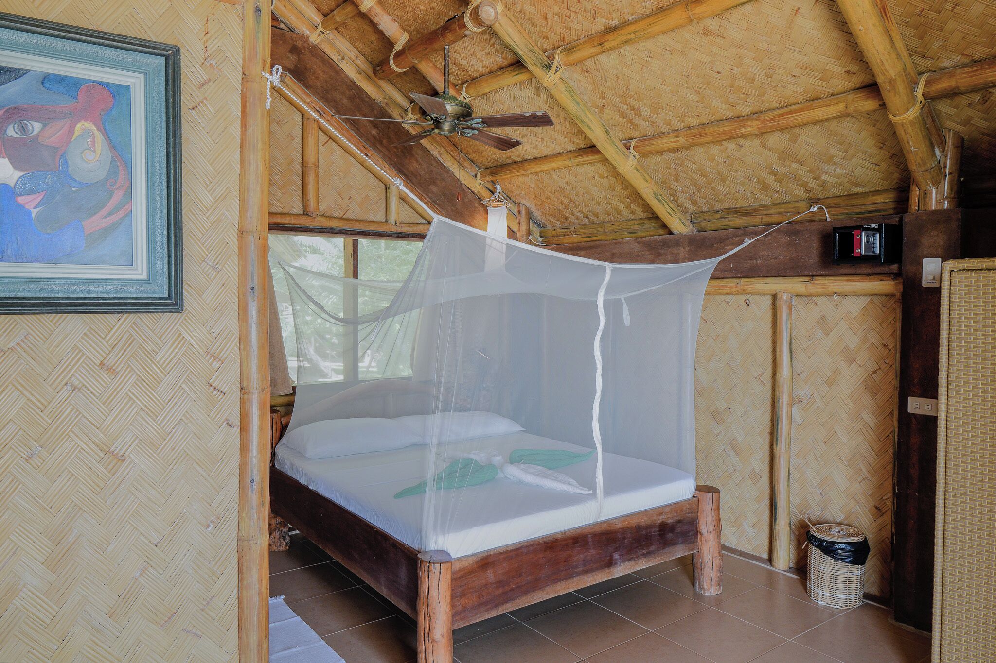 A bed with a mosquito net, wooden beams, a ceiling fan, and a framed picture on the wall.