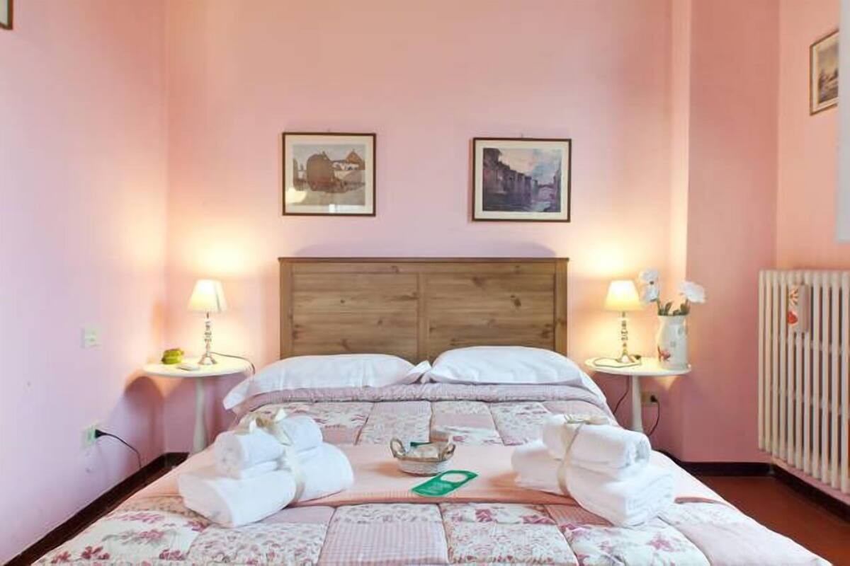 Een slaapkamer met een houten hoofdeinde, twee nachtkastjes met lampen, een bloemenbedekking en twee ingelijste foto’s aan de muur.