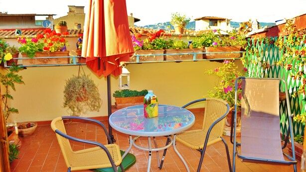 Une table ronde en verre, des chaises en métal, un parasol rouge et des plantes en pot sur une terrasse.