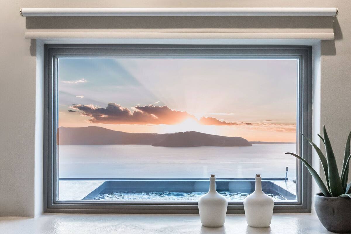 A window with a view of a sunset over water, two white vases, and a potted plant on a windowsill.