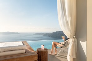 A person relaxing on a chair by a pool with a view of the sea and mountains. - West East Suites (Santorini)