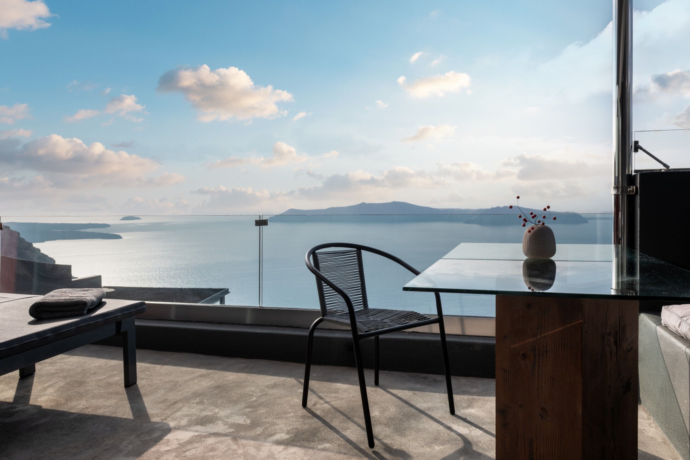 Un balcón con vistas al mar, una mesa de vidrio, una silla de metal y un armario de madera.