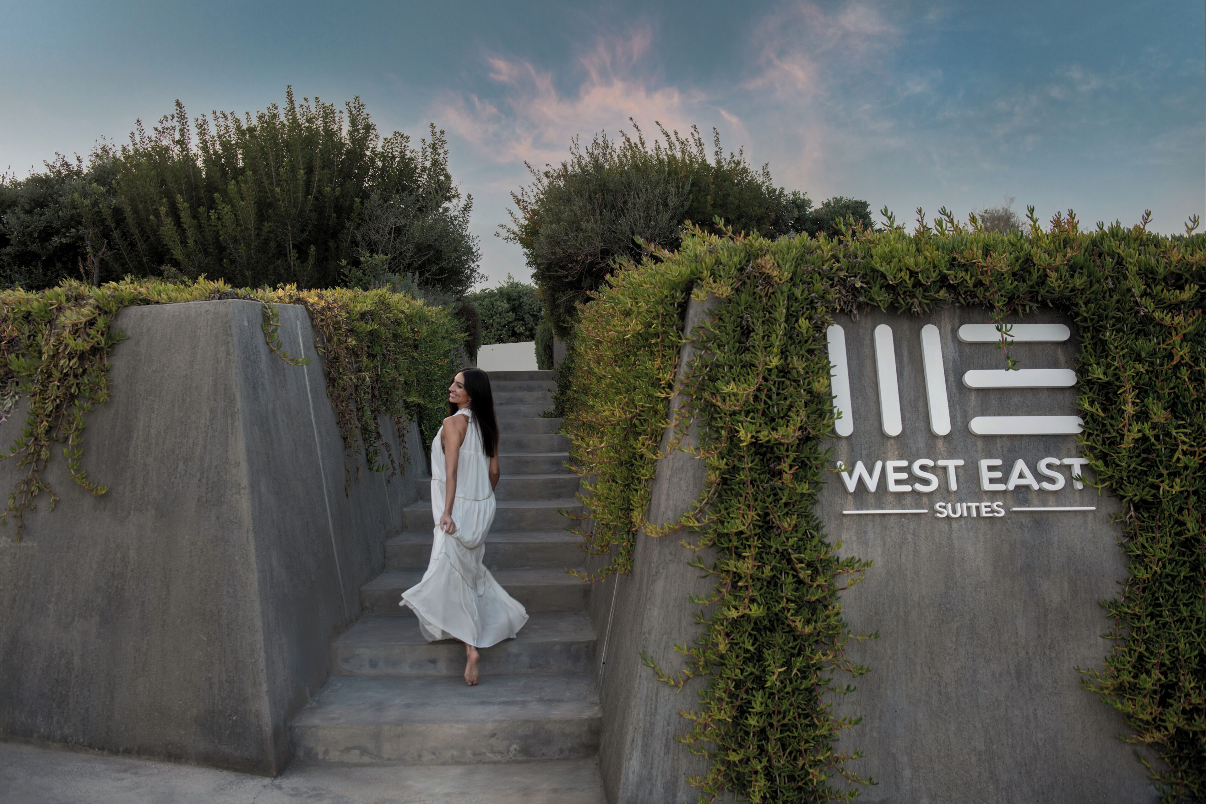 A woman descending concrete steps with a sign reading 'WEST EAST SUITES' amidst greenery.