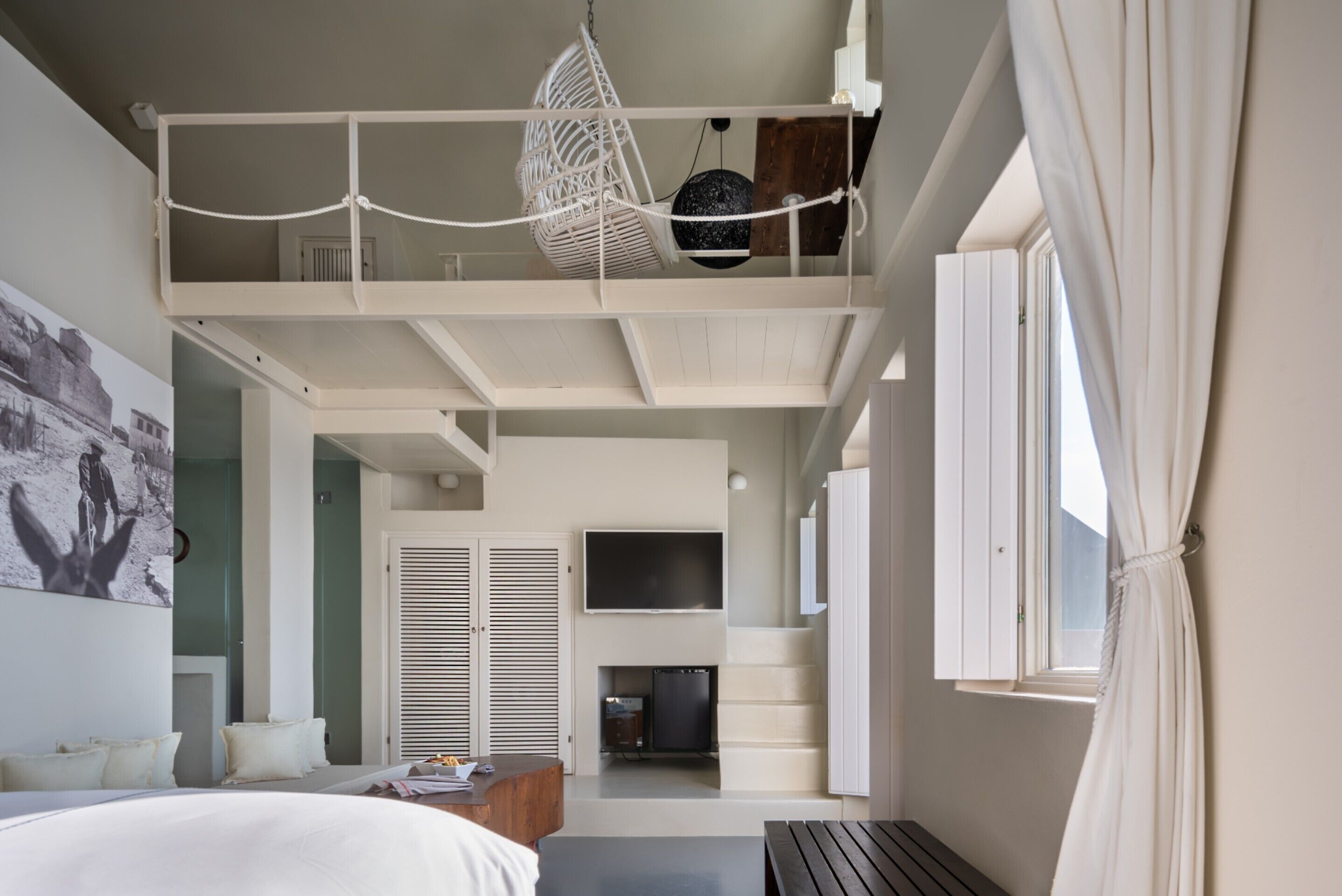 A modern hotel room with a loft bed, a flat-screen TV, a seating area with a table, and a large window.