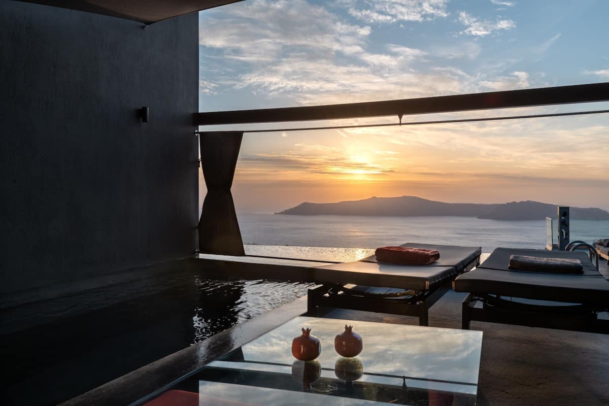 A modern room with a large window, a glass table with two pomegranates, a red cushion, and a view of the sunset over the sea.