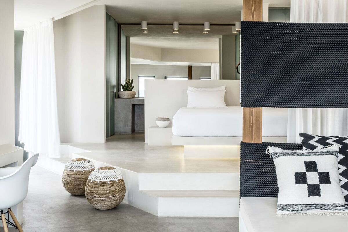 A modern hotel room with a minimalist design, featuring a large bed, a seating area with a sofa and cushions, and a small table with a plant.