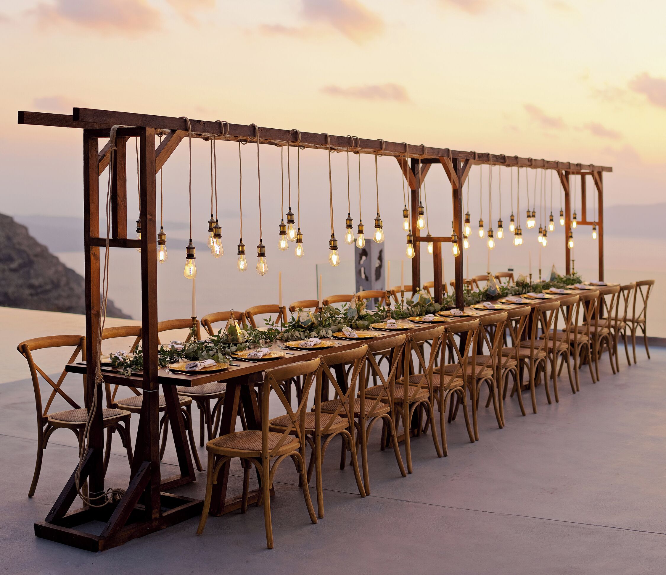 A long wooden table set for a meal with hanging light bulbs above, surrounded by wicker chairs.