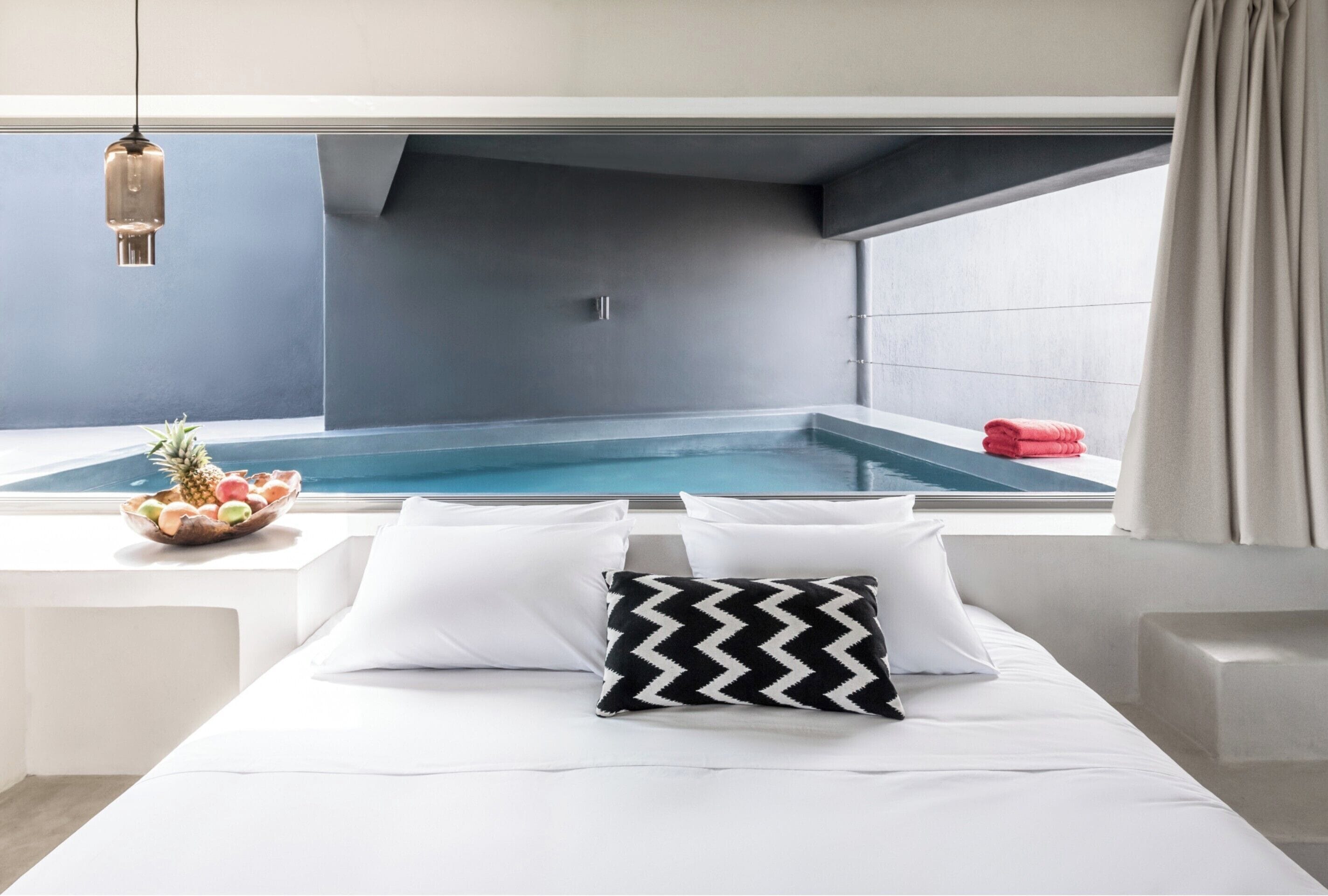 Ein modernes Schlafzimmer mit Blick auf einen Swimmingpool, ein Bett mit weißer Bettwäsche, ein Schwarz-Weiß-Kissen und eine Obstschale auf der Fensterbank.