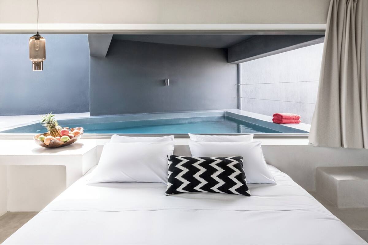 A modern bedroom with a view of a swimming pool, a bed with white bedding, a black and white pillow, and a fruit bowl on the windowsill.
