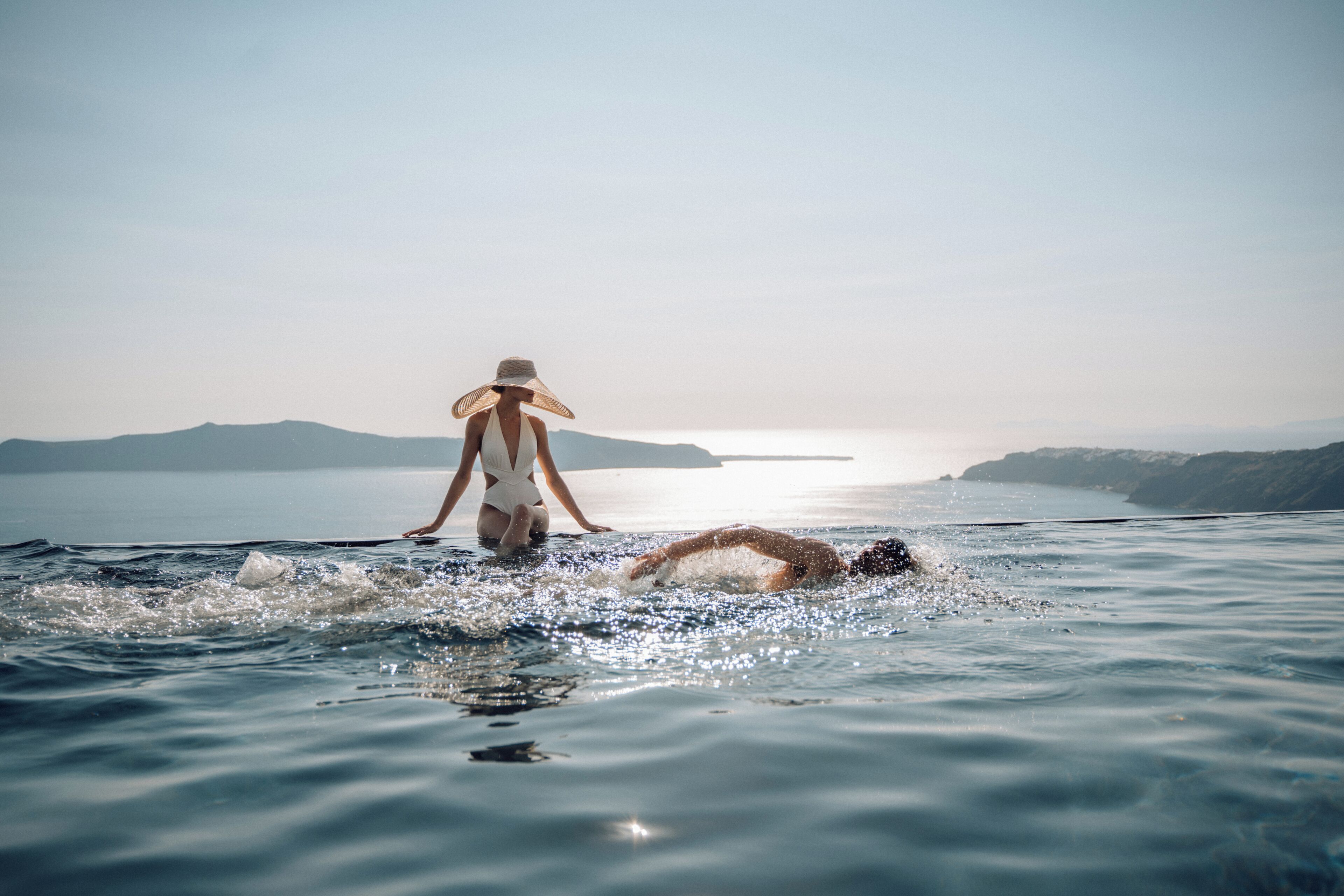 A person in a swimsuit and hat standing in calm water with a distant shoreline and mountains.