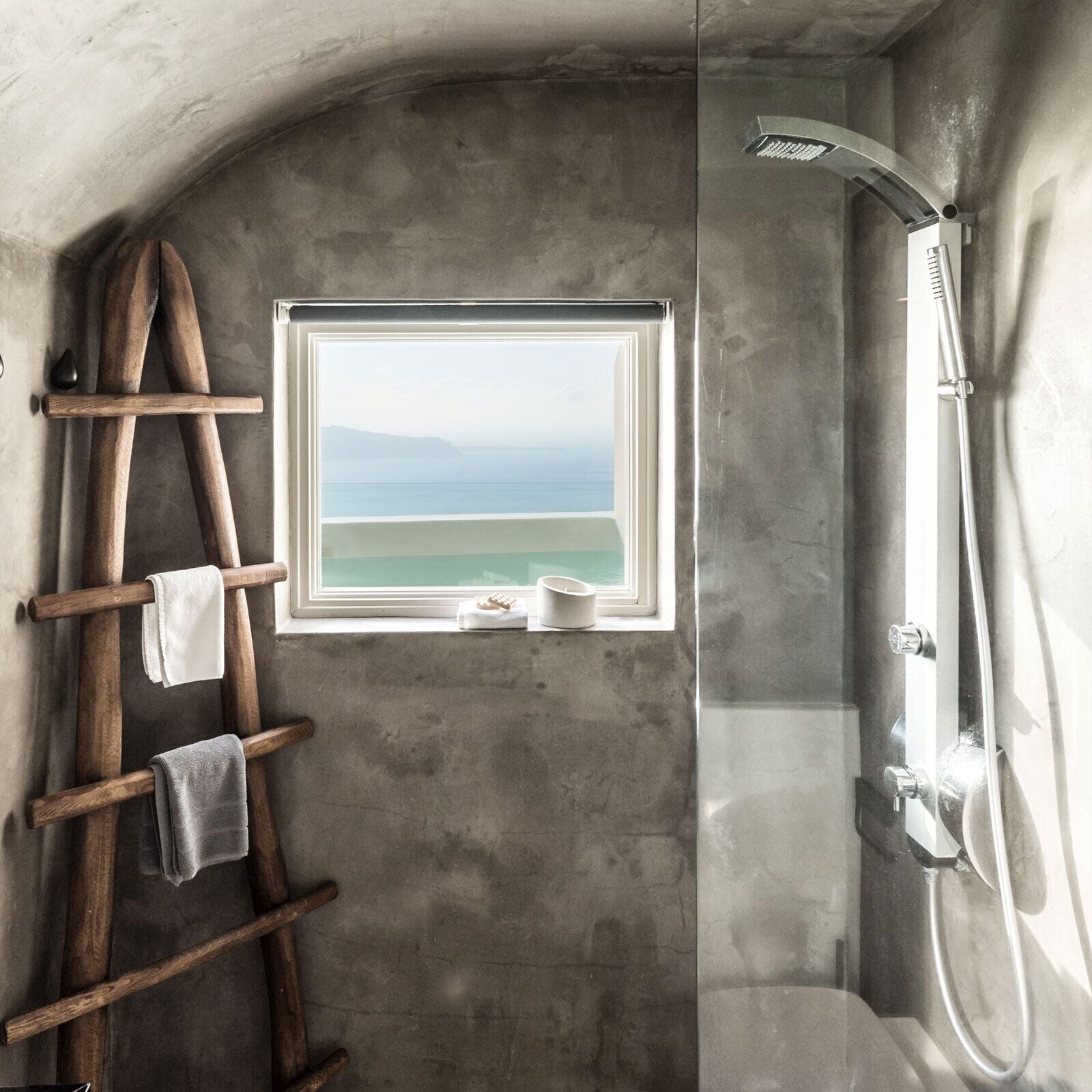 A bathroom with a concrete wall, a wooden ladder, a window with a view of the sea, and a shower.