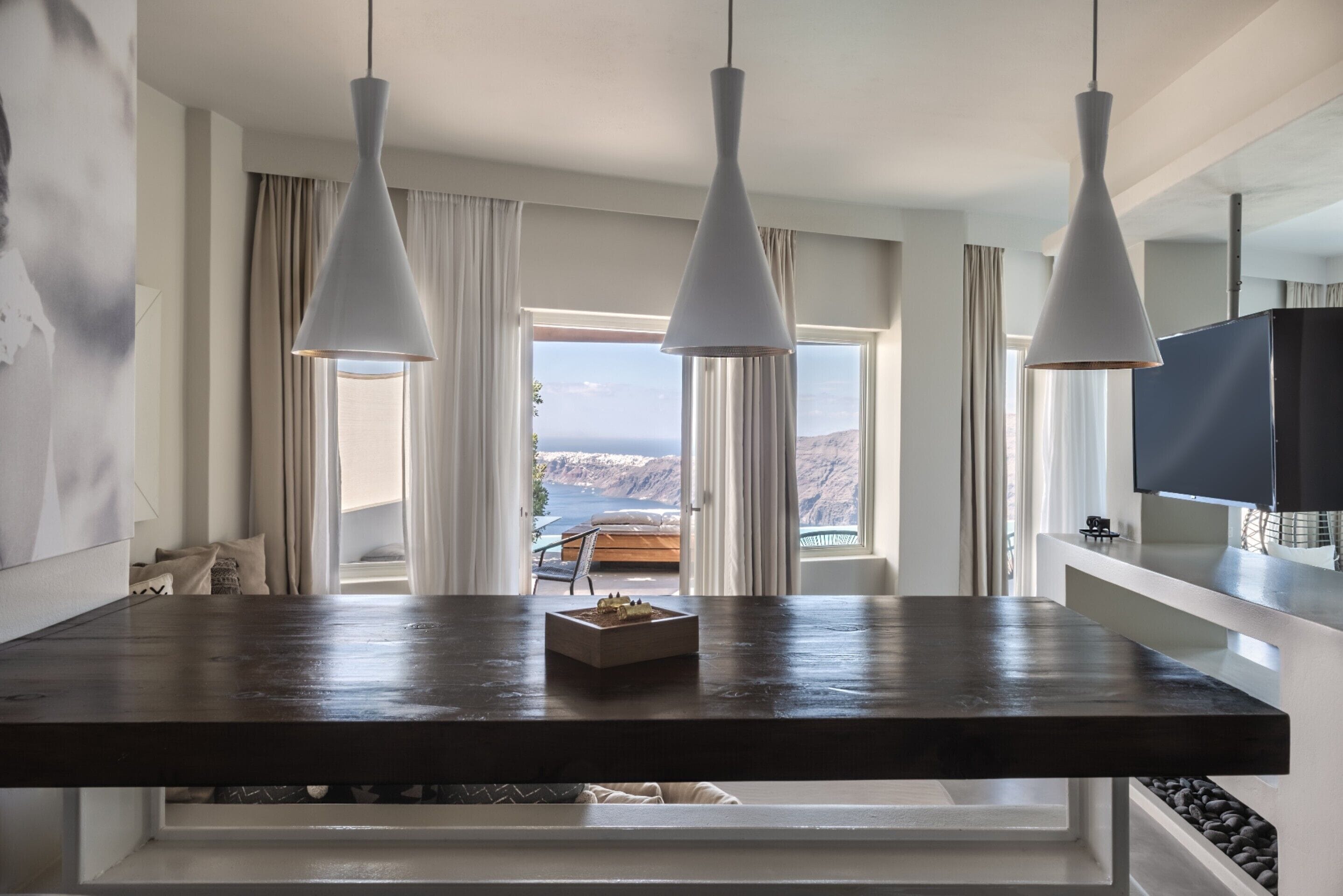 Comedor con una mesa de madera oscura, tres lámparas colgantes blancas y vistas al mar a través de amplios ventanales.
