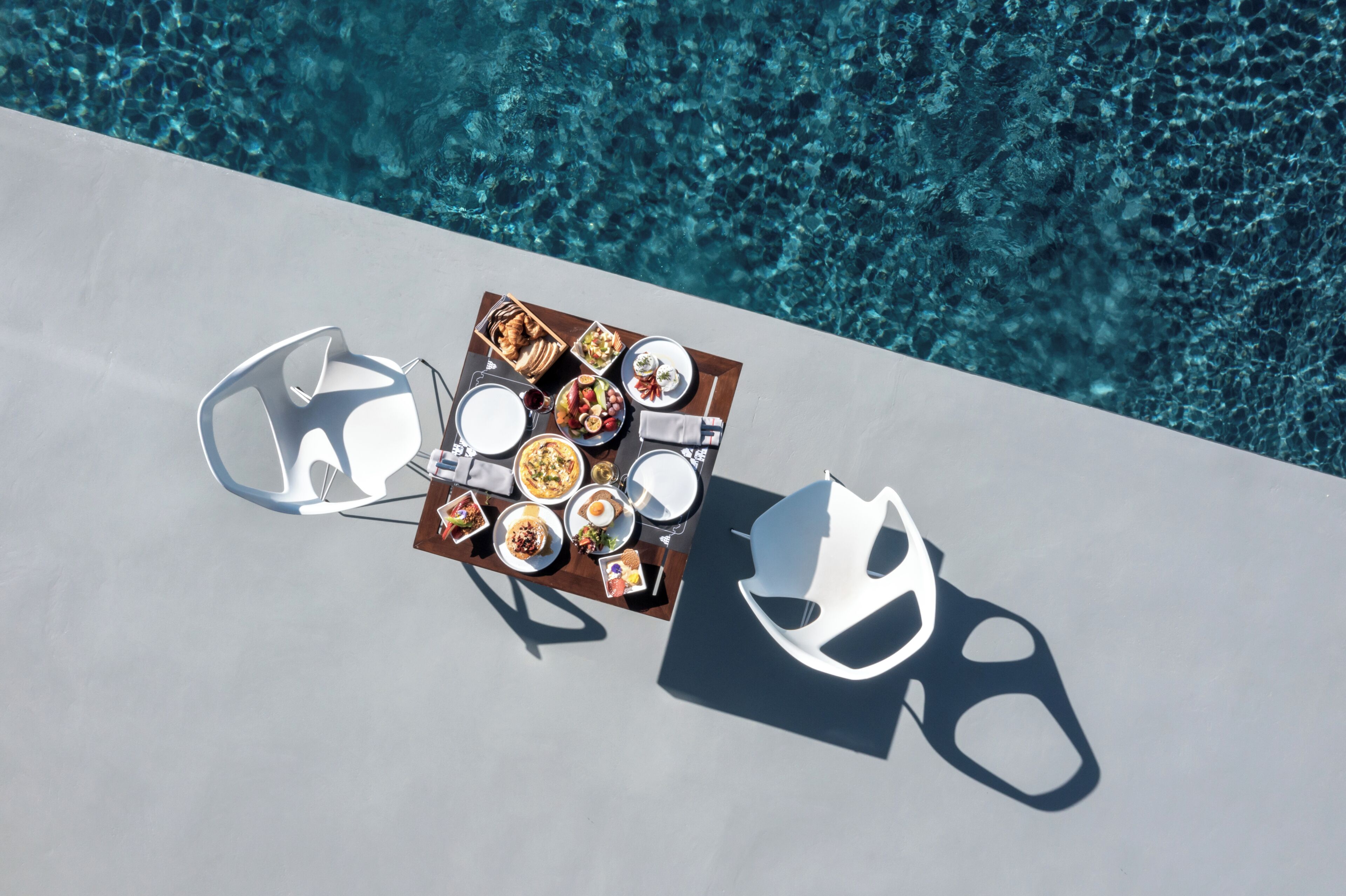 Vue aérienne d’une table à manger avec différents plats, deux chaises blanches et un décor de bord de piscine.
