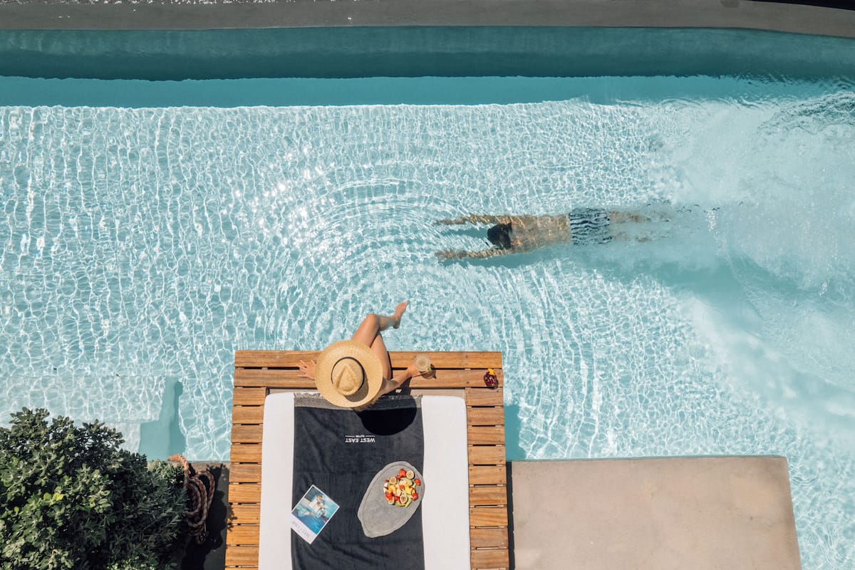 A swimming pool with clear water, a wooden deck, lounge chairs, and a towel with a book and fruit.