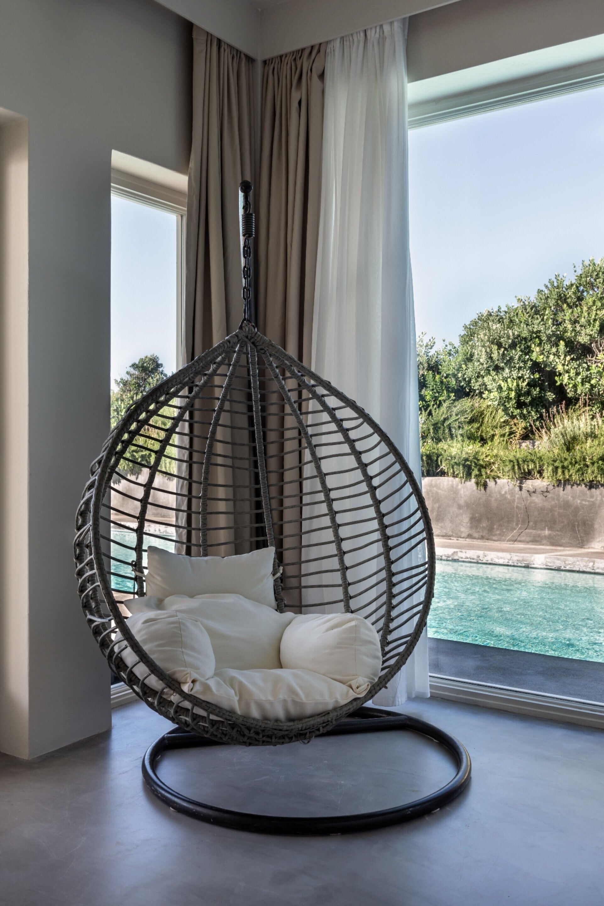 A hanging chair with cushions by a window overlooking a pool.