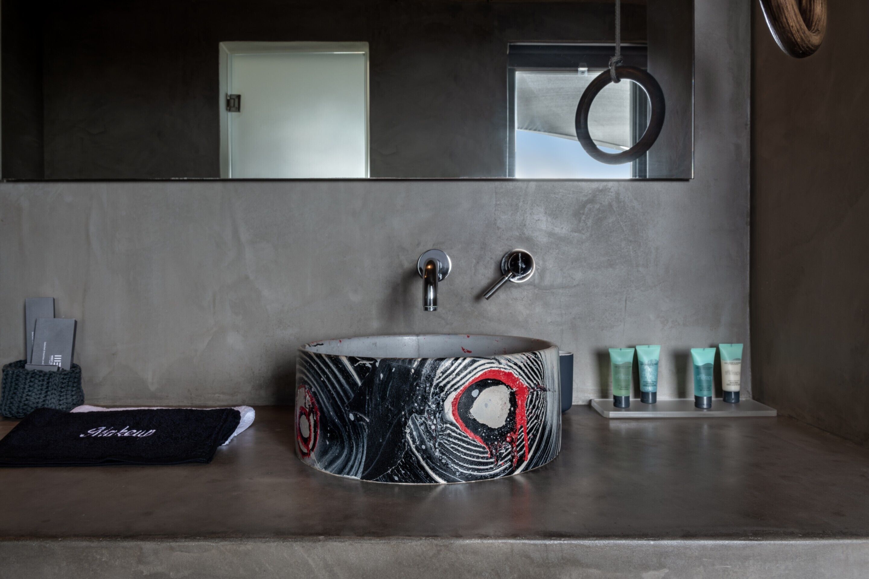 A modern bathroom with a round sink, two faucets, and a mirror above. There are toiletries on the counter and a towel on the floor.