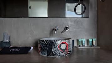 A modern bathroom with a round sink, two faucets, and a mirror above. There are toiletries on the counter and a towel on the floor.