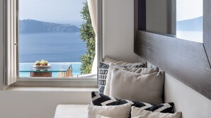 Habitación con un amplio ventanal que ofrece vistas al mar y a un paisaje montañoso. Hay un sofá con cojines y una mesita con una planta.