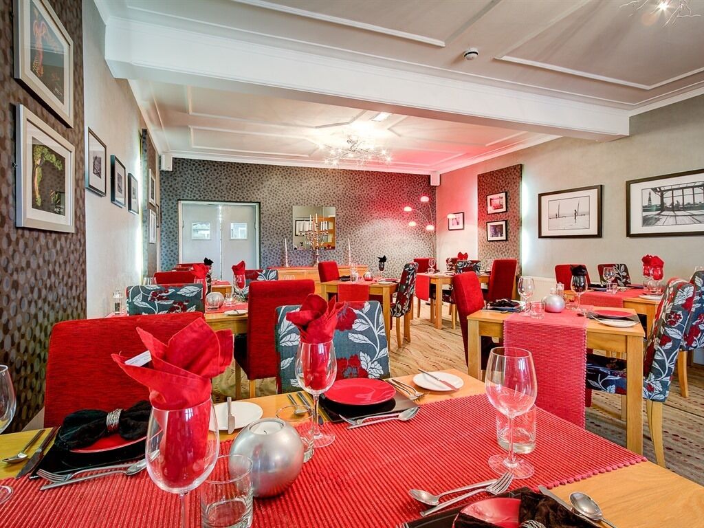 A restaurant with red and blue chairs, tables set with red tablecloths, and framed pictures on the walls.