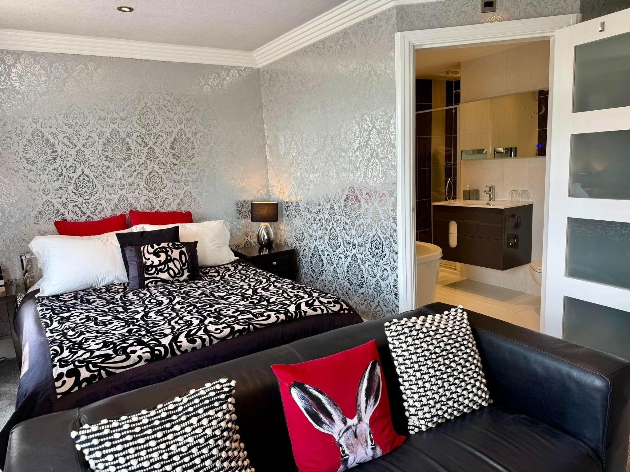 A bedroom with a patterned bedspread, a red and white pillow, a black and white patterned pillow, a black leather sofa with a red pillow, and a bathroom visible through an open door.