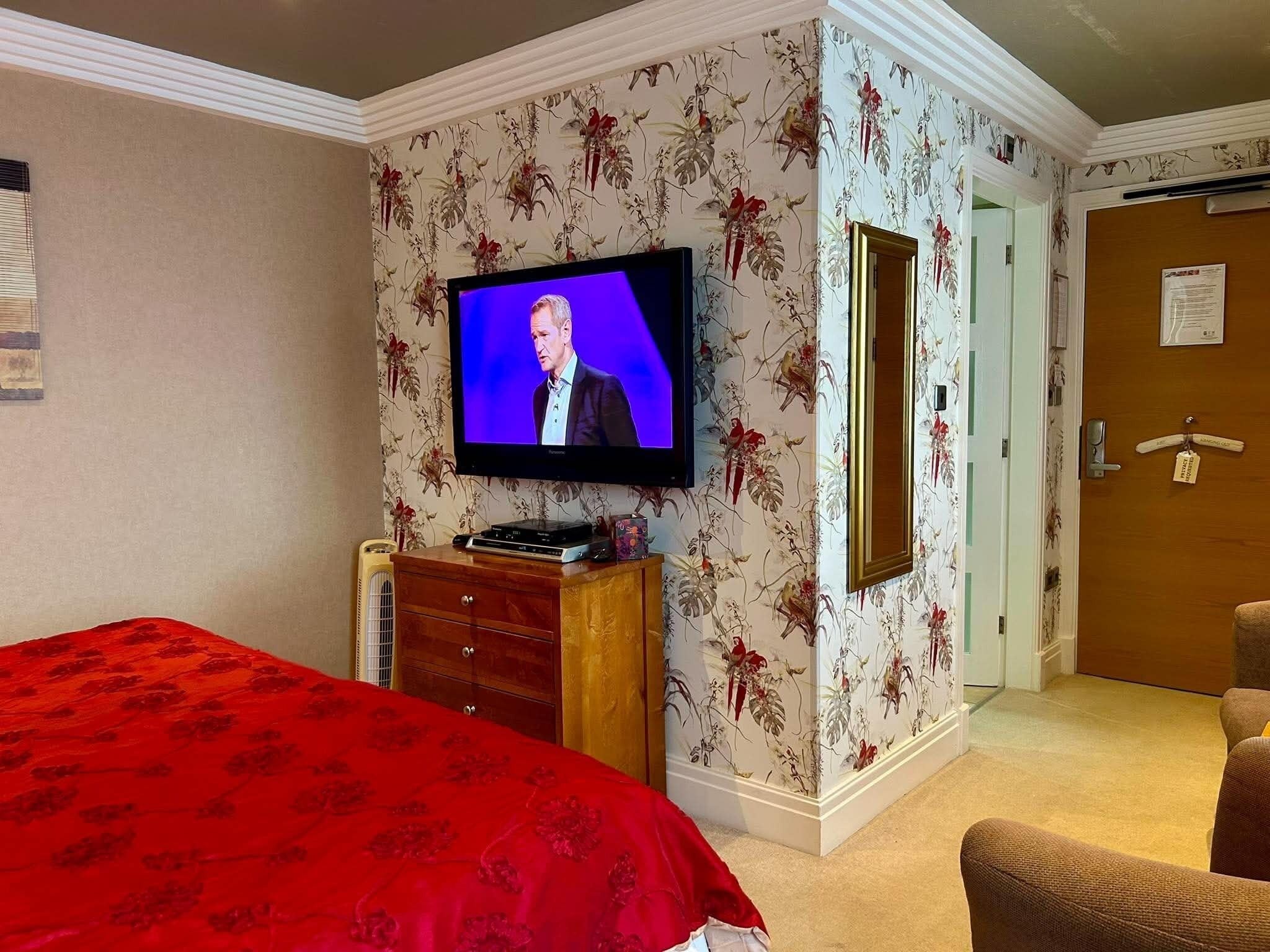 Ein Hotelzimmer mit Fernseher an der Wand, einem Bett mit roter Bettdecke, einer Holzkommode und einer Tür.