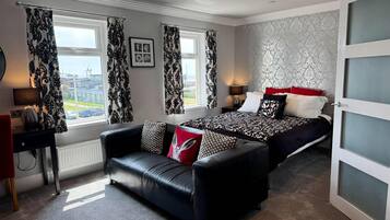A modern bedroom with a black leather sofa, a bed with patterned bedding, and a view of the city through large windows.