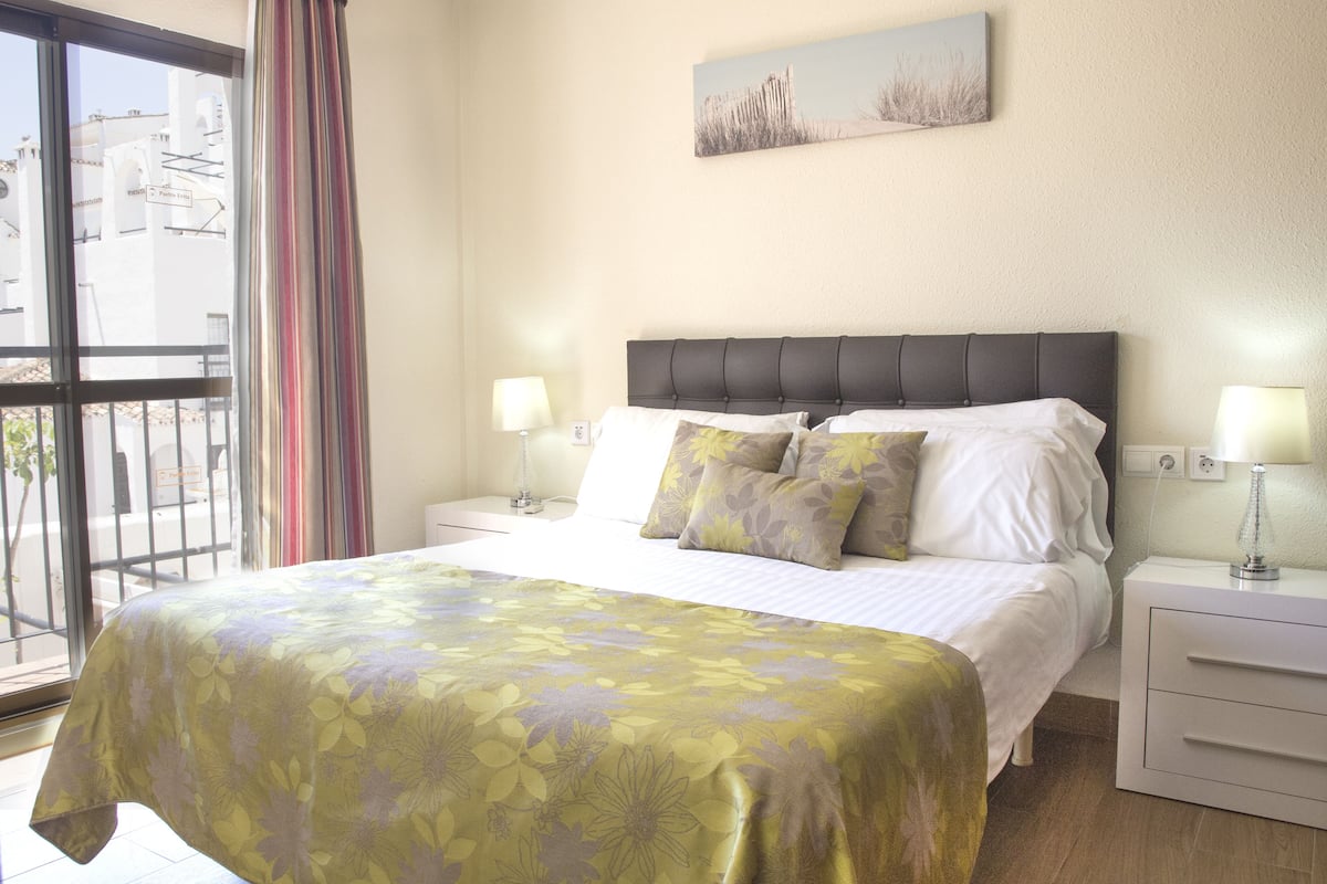 Een hotelkamer met een bed, twee nachtkastjes met lampen, een raam met gordijnen en een schilderij aan de muur.