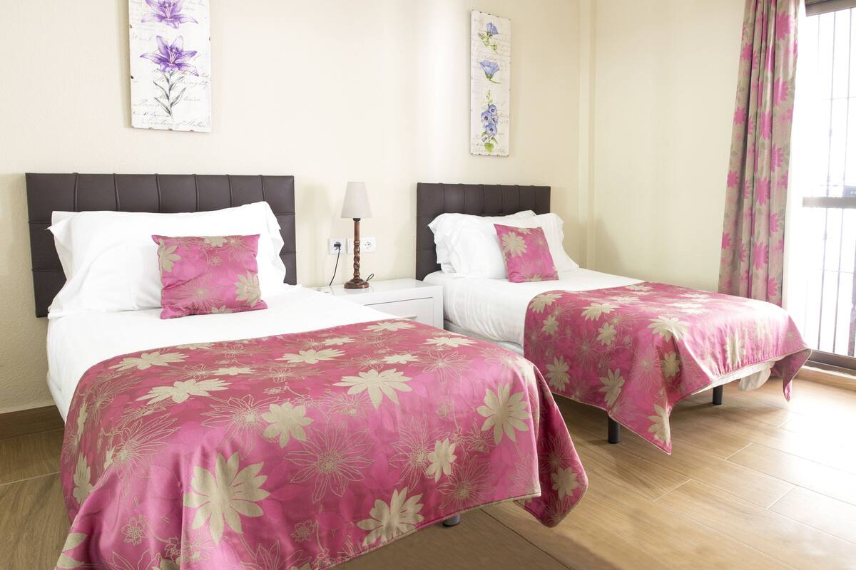 Een hotelkamer met twee bedden, elk voorzien van een roze bloemenlaken, een nachtkastje met een lamp en wanddecoratie boven de bedden.