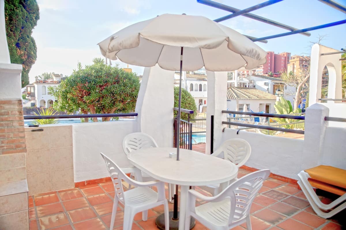 Een terras met een witte tafel en stoelen, een parasol en uitzicht op gebouwen en bomen.