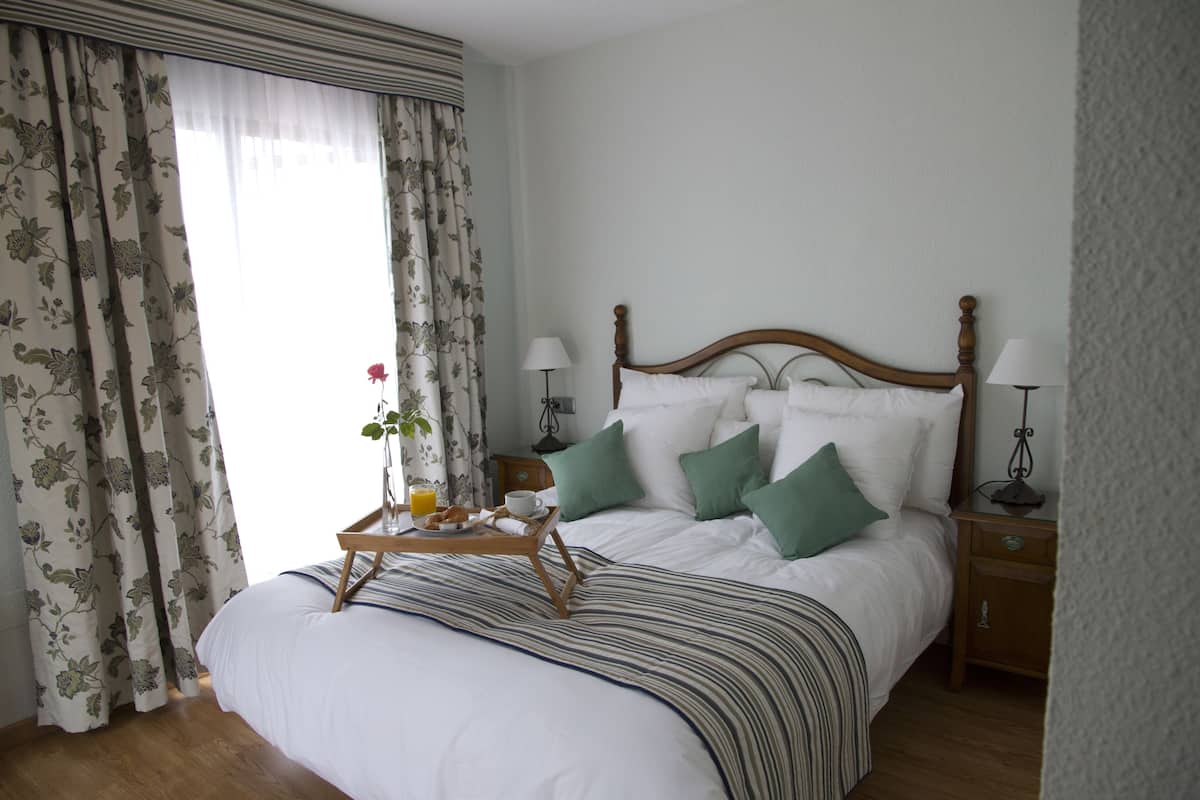 A neatly made bed with a tray of breakfast, a bedside table with a lamp, and a window with curtains.