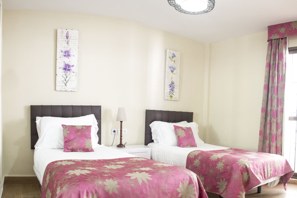 Een hotelkamer met twee bedden, elk met een roze bloemenbedekking, witte kussens en een donker hoofdbord. Tussen de bedden staat een nachtkastje met een lamp, en aan de muur hangt een bloemistisch kunstwerk.