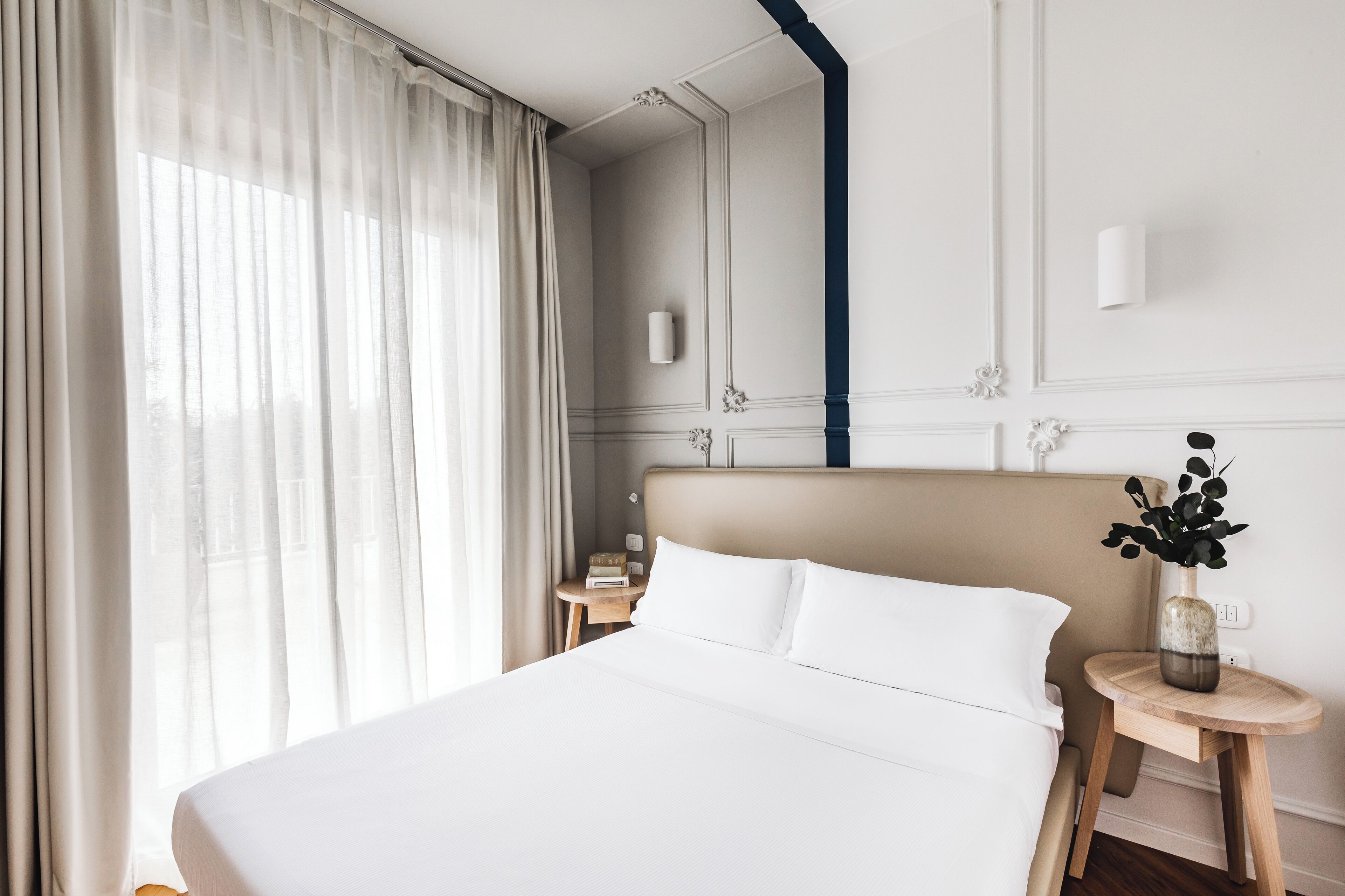 Habitación de hotel con una cama grande, una mesita de noche, una pequeña mesa con un florero y una ventana con cortinas translúcidas.