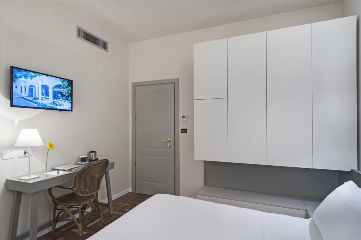 Ein Hotelzimmer mit einem Bett, einem Schreibtisch mit Lampe, einem Sessel, einem an der Wand befestigten Fernseher und einem Kleiderschrank.