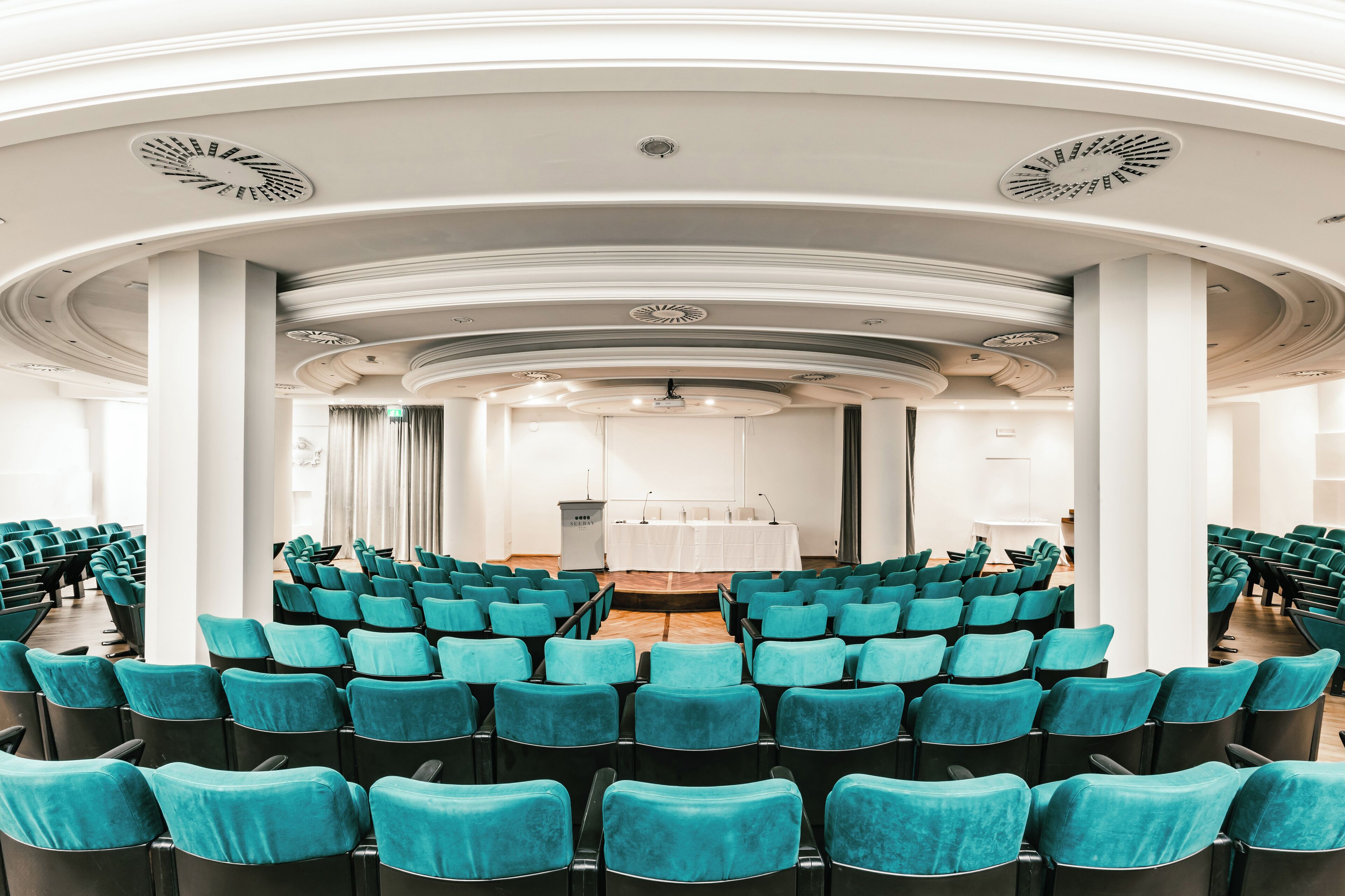 Ein Konferenzraum mit Reihen aus türkisfarbenen Stühlen, weißen Säulen und einer Bühne mit Rednerpult.