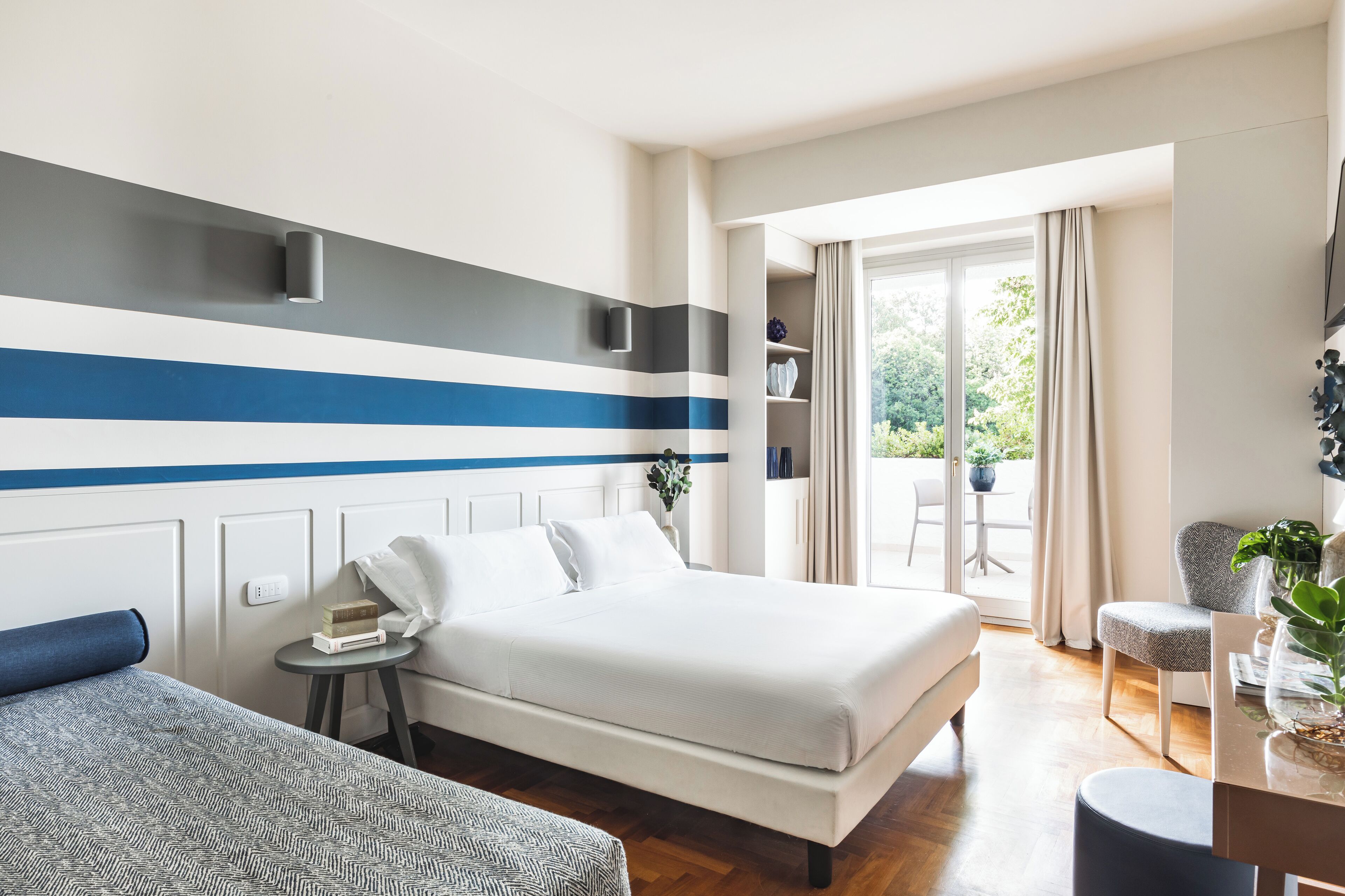 Ett modernt hotellrum med en stor säng, ett litet sidobord, en soffa, en balkong med utsikt över grönområden och en vägg med horisontella ränder.