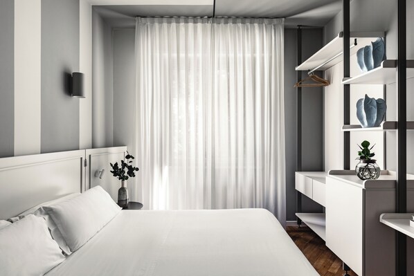 A modern bedroom with a large bed, a bedside table with a vase, a wardrobe with shelves, and a window with sheer curtains.
