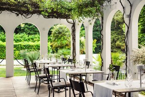 An outdoor dining area with tables and chairs, surrounded by white arches and greenery.