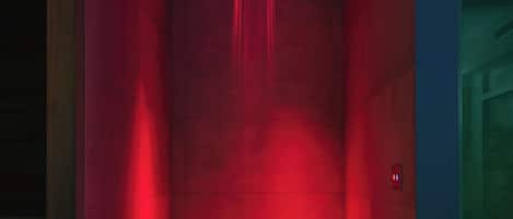A red-lit elevator with a modern design, featuring a control panel on the wall and a visible emergency exit sign.
