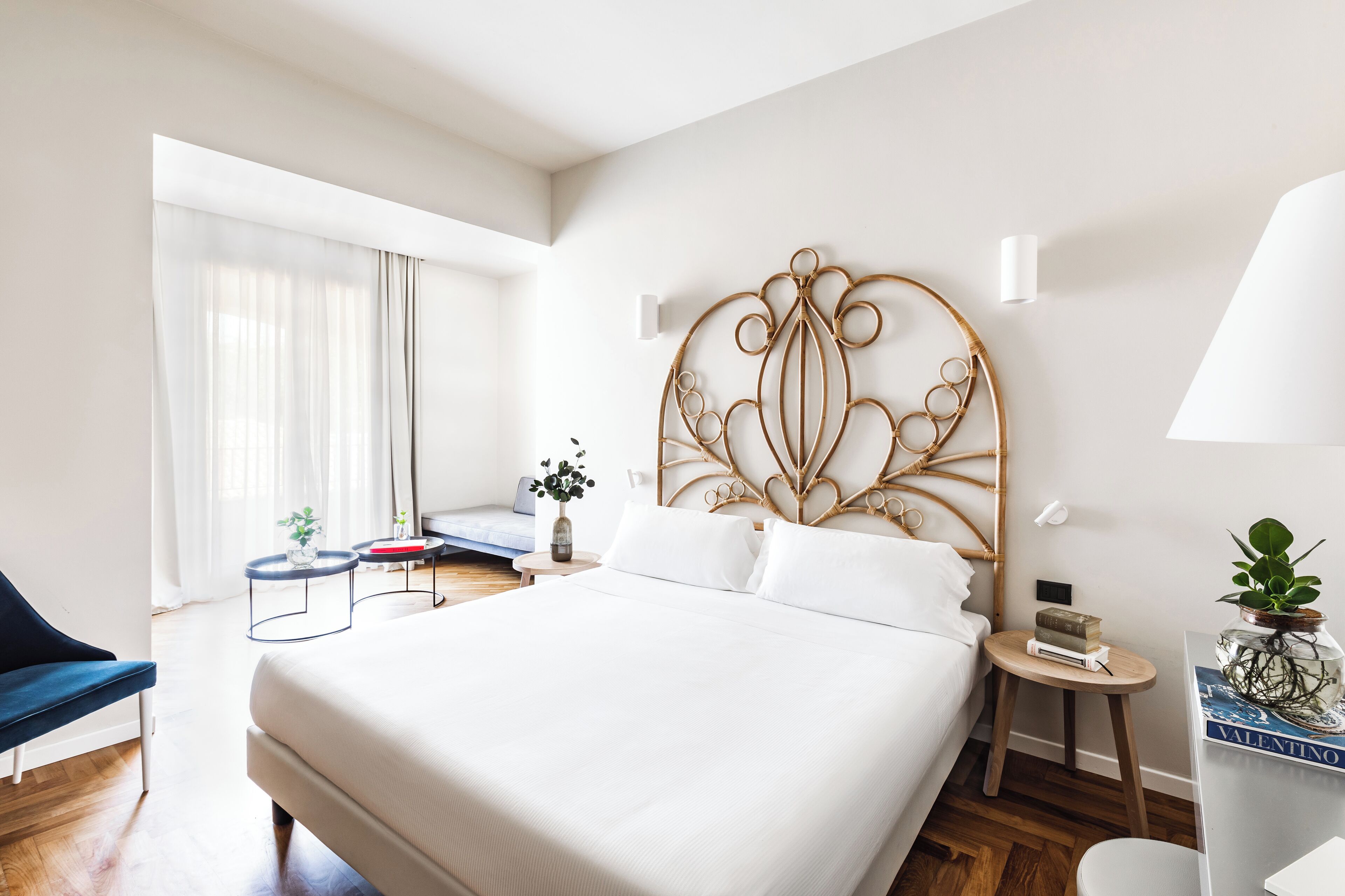 A hotel room with a large bed, a bedside table with a lamp, a small side table with a book and a plant, a chair, and a window with sheer curtains.