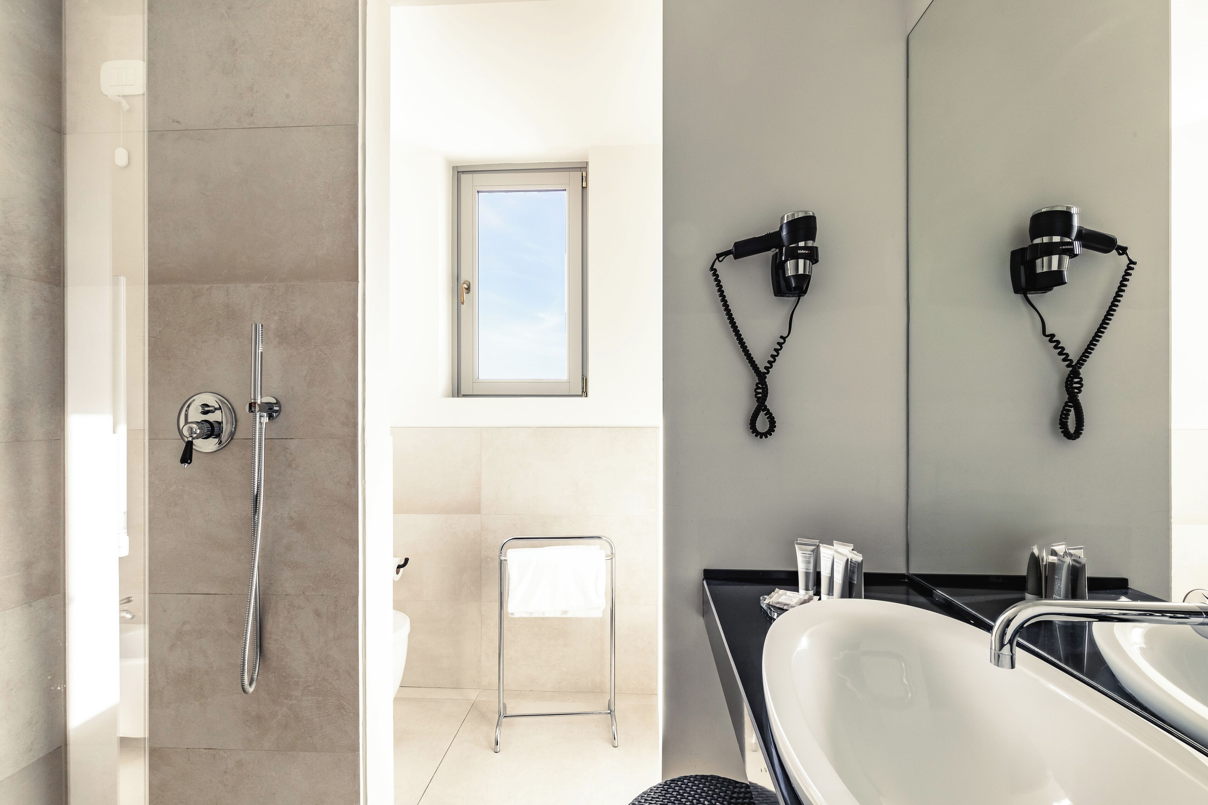 Een moderne badkamer met een douche, een klein krukje, een wastafel met spiegel en twee haardroger bevestigd aan de muur.