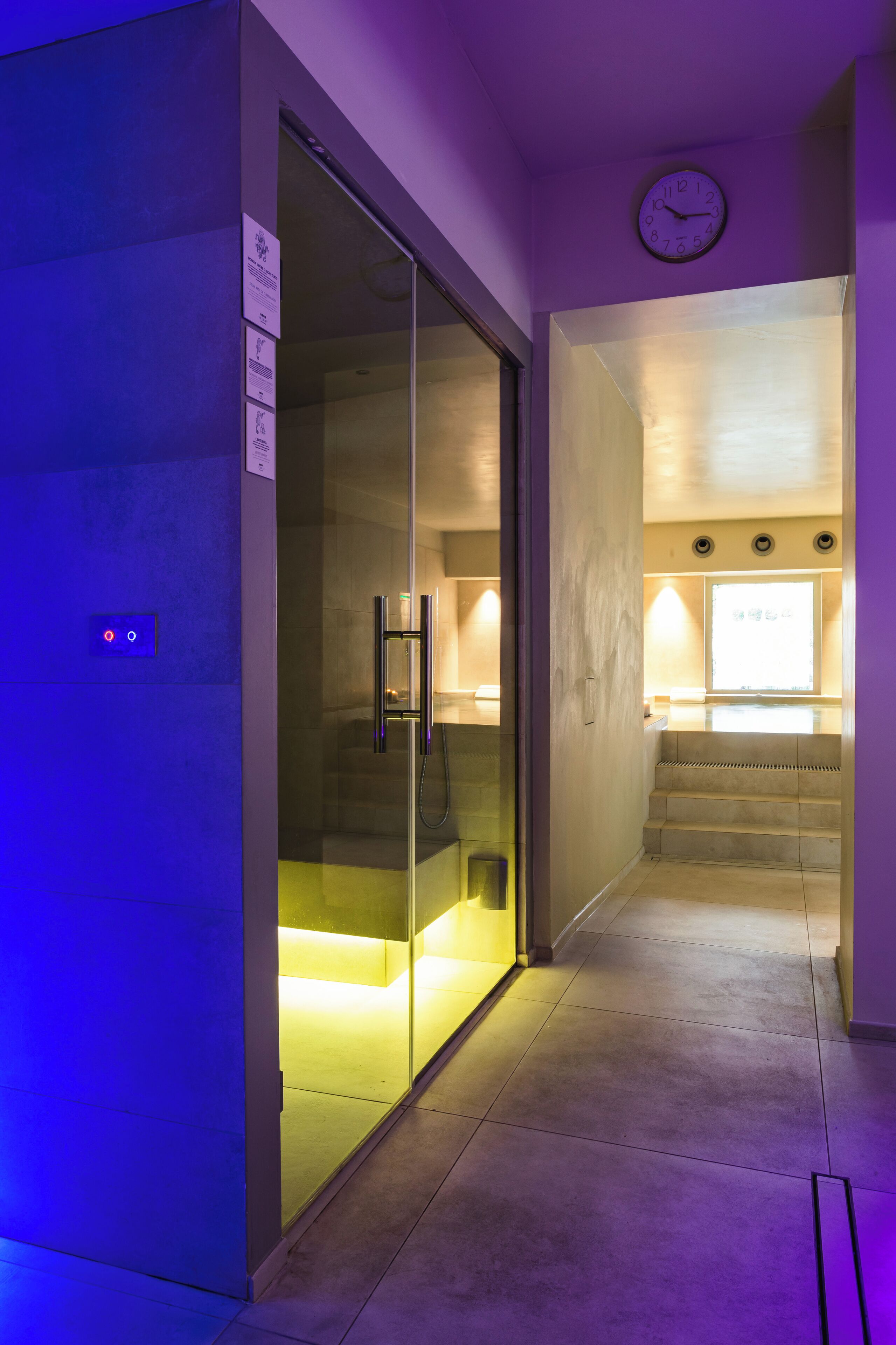 A hallway with a glass door leading to a bathroom, a clock on the wall, and a staircase.