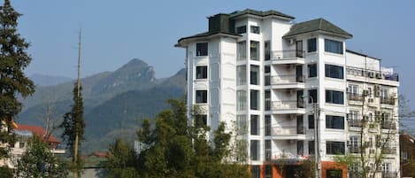 Un edificio de varios pisos con balcones, rodeado de vegetación y montañas al fondo.