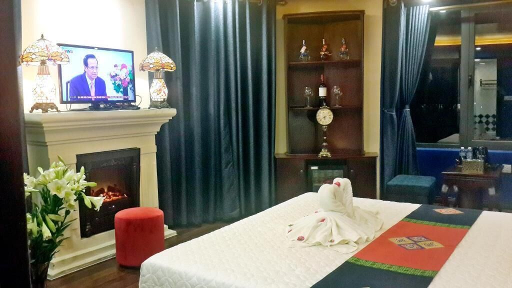 A hotel room with a fireplace, a bed with a white bedspread, a red ottoman, a television on a mantelpiece, and a window with curtains.