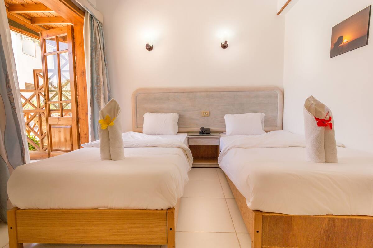 A hotel room with two beds, wooden headboards, white bedding, and a small bedside table with a lamp.