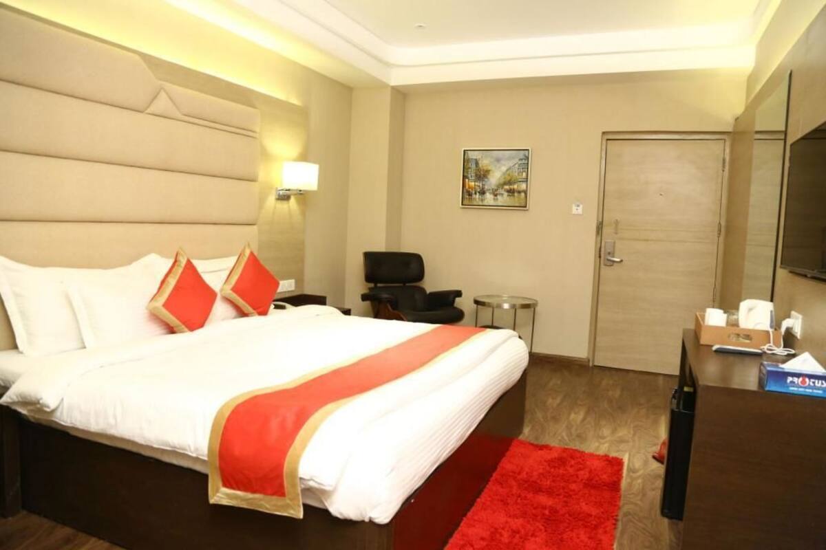 Quarto de hotel com uma cama grande, um cobertor vermelho, uma escrivaninha pequena com cadeira, uma televisão e uma porta.